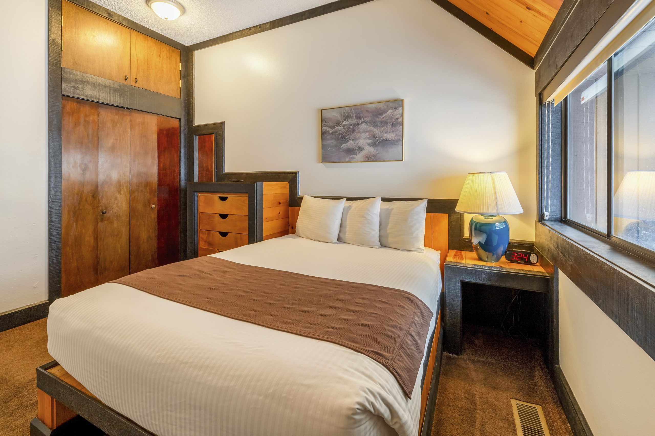 A hotel room with a large bed, a nightstand with a lamp, a painting on the wall, and a window with a view.