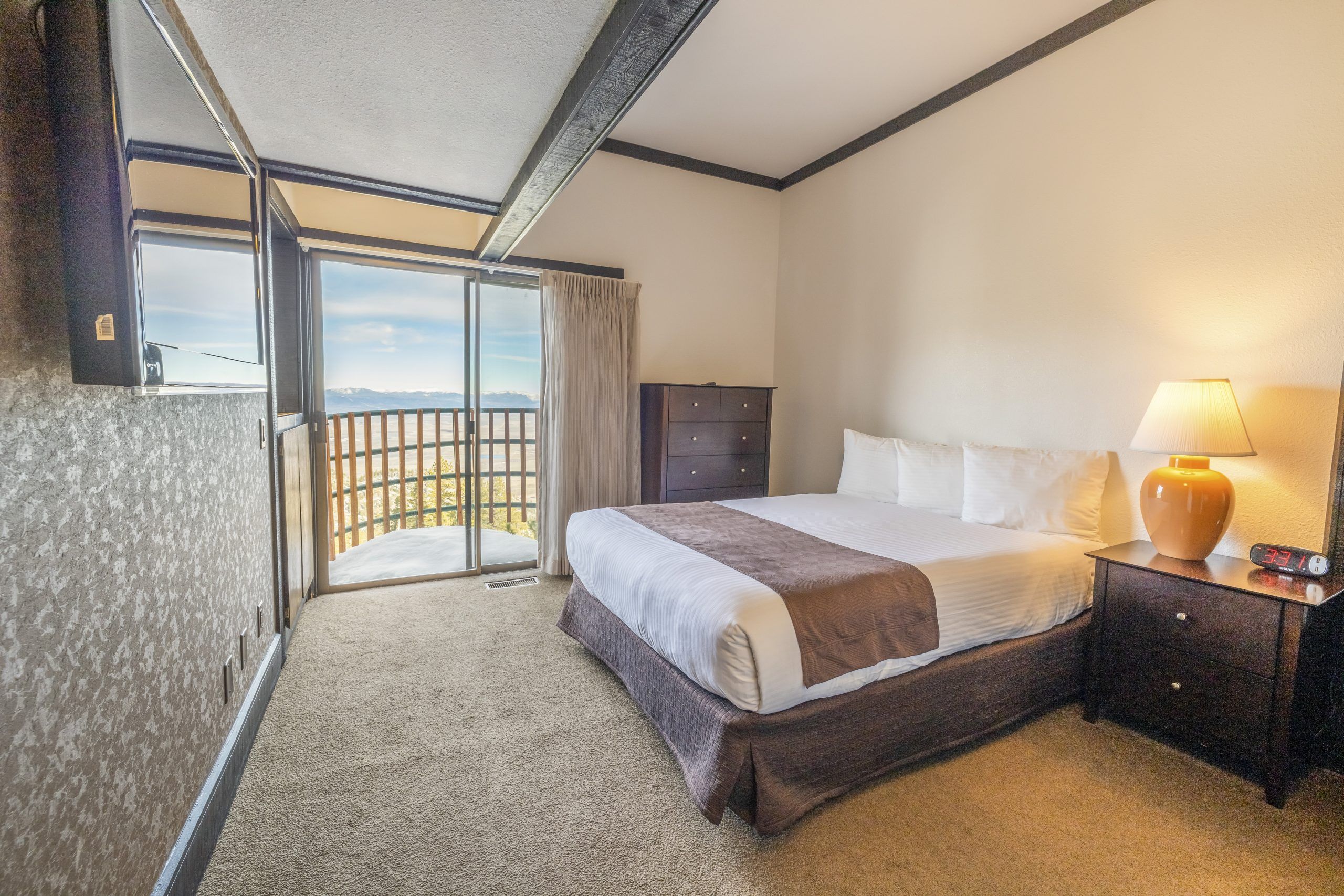 A hotel room with a bed, a nightstand, a lamp, a digital clock, and a view of a snowy landscape through a sliding door.