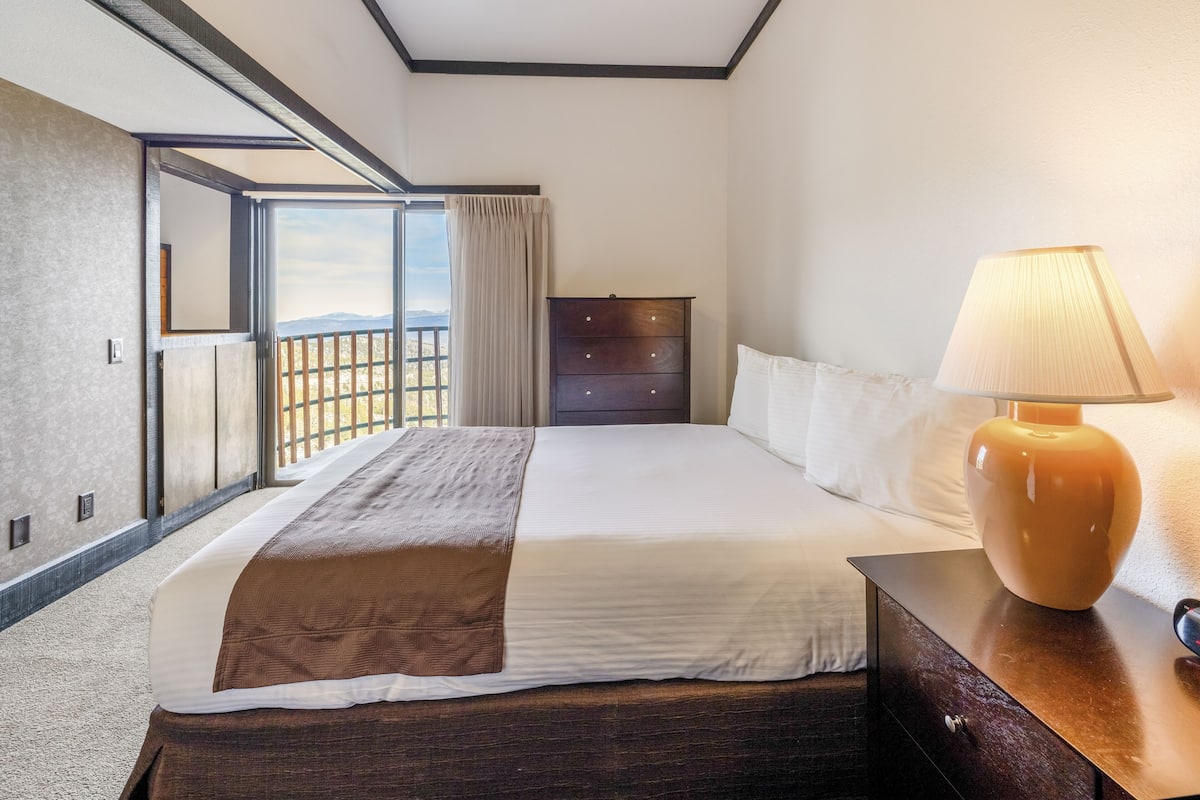 A hotel room with a large bed, a nightstand, a lamp, a dresser, and a sliding glass door leading to a balcony.