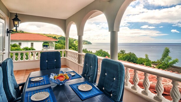 A balcony with a table set for dining, overlooking a scenic view of the sea and greenery. - Caribbean Sea View Holiday Apartments (Mero)