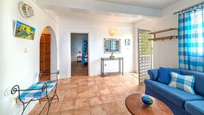A living room with a blue sofa, a coffee table, and a blue checkered chair. There is a door leading to another room and a window with blue curtains. - Caribbean Sea View Holiday Apartments (Mero)