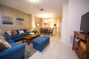 A living room with a blue sofa, a coffee table, and a TV. There is a dining area with a table and chairs.