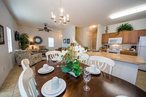 A dining area with a wooden table set for four, surrounded by white chairs. A chandelier hangs from the ceiling, and a ceiling fan is visible. The room has a modern kitchen in the background with wooden cabinets and a microwave.