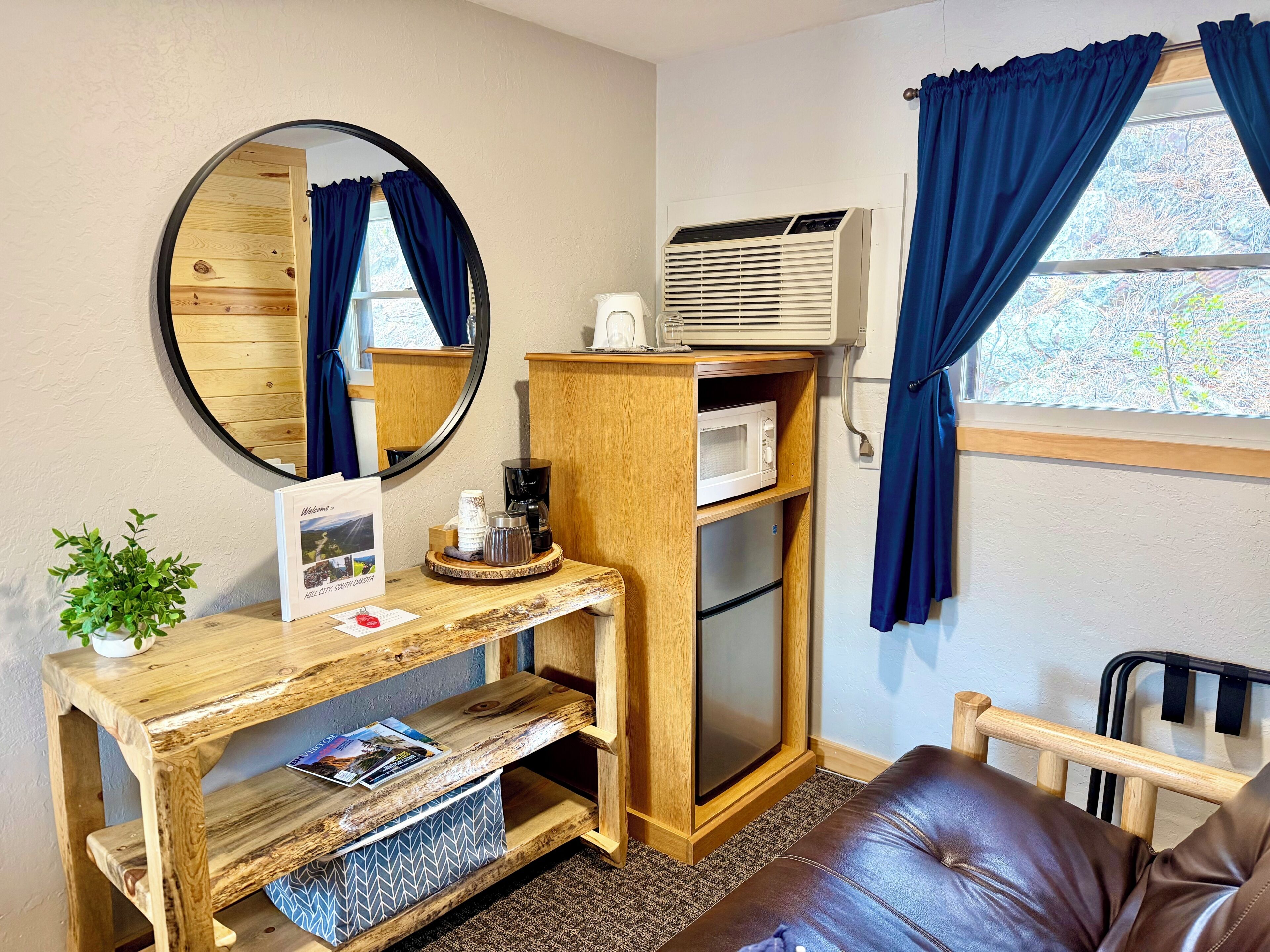 A room with a wooden desk, a mirror, a microwave, a coffee maker, a small plant, and a leather couch.