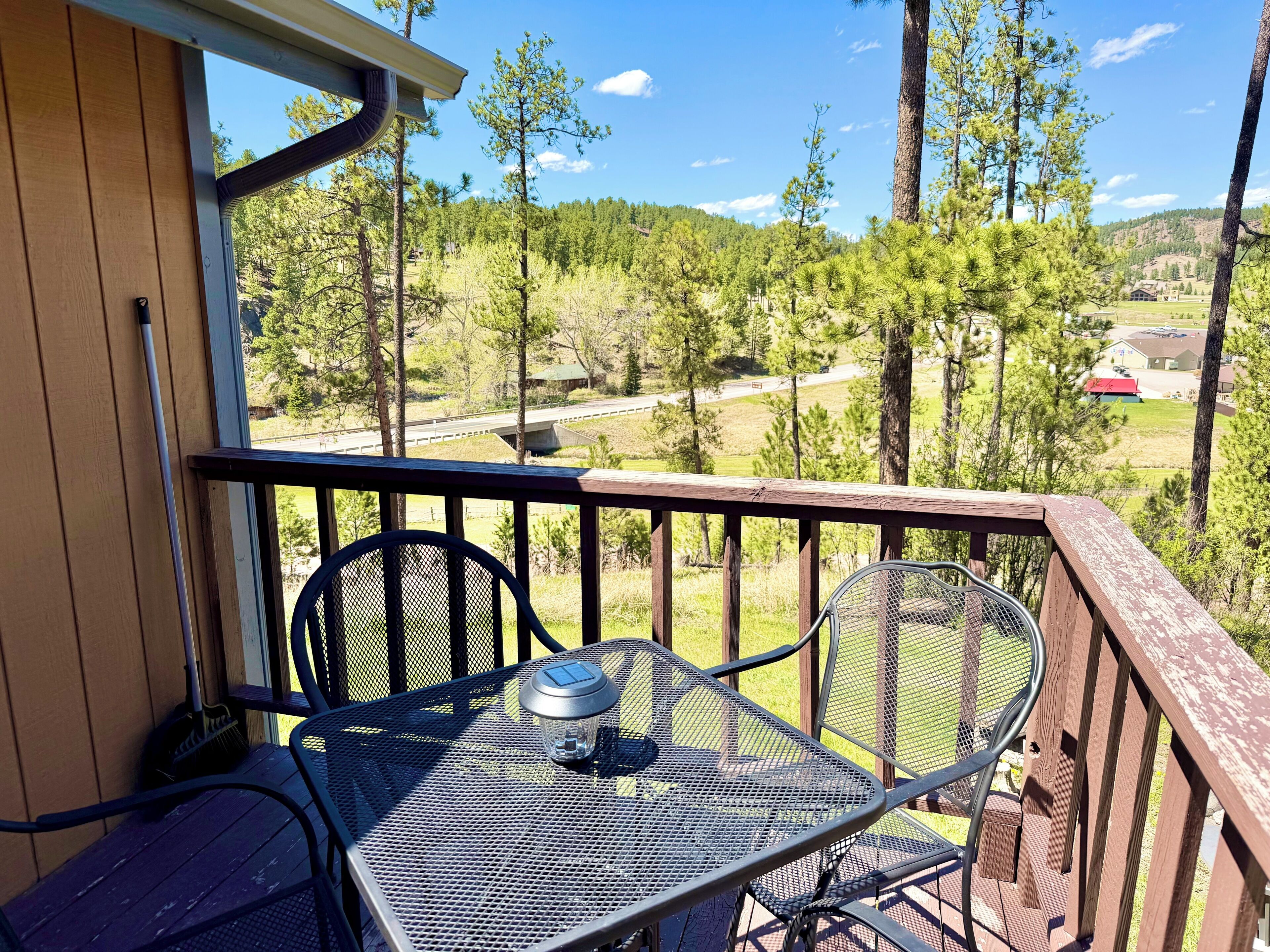 Ein Balkon mit einem Metalltisch und -stühlen, der auf eine bewaldete Landschaft mit blauem Himmel blickt.