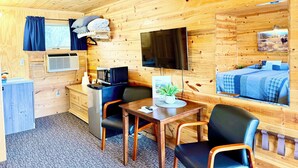 A wooden cabin interior with a bed, a TV, a microwave, a table with a chair, and a window with curtains. - Mountain View Lodge & Cabins (Rapid City)