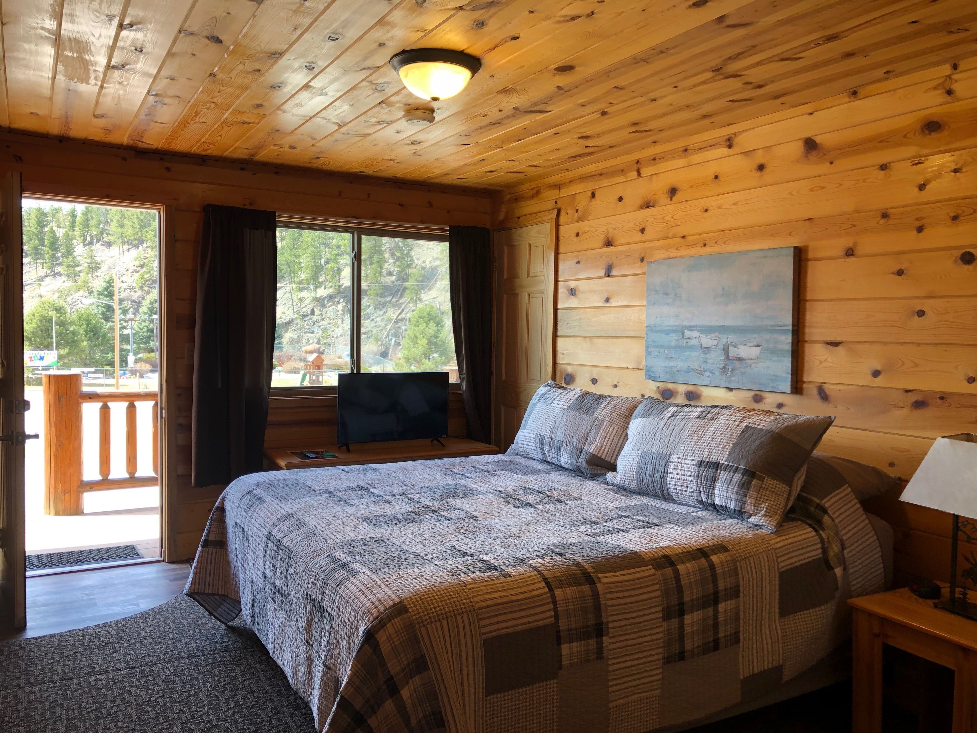 A wooden cabin bedroom with a bed, a painting on the wall, and a balcony with a view of trees and a mountain.