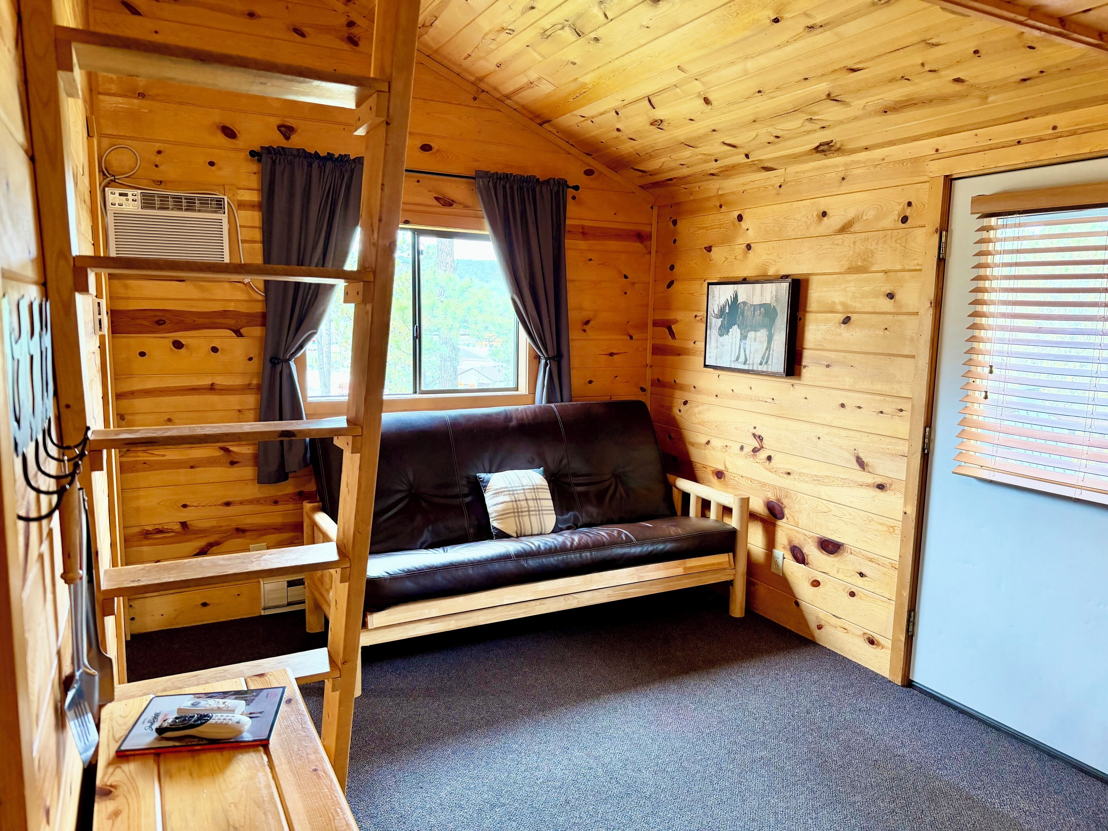 A cozy cabin interior with a wooden interior, a sofa, a ladder, a window with curtains, and a picture on the wall.