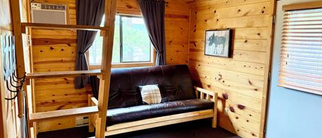 A cozy cabin interior with a wooden interior, a sofa, a ladder, a window with curtains, and a picture on the wall.