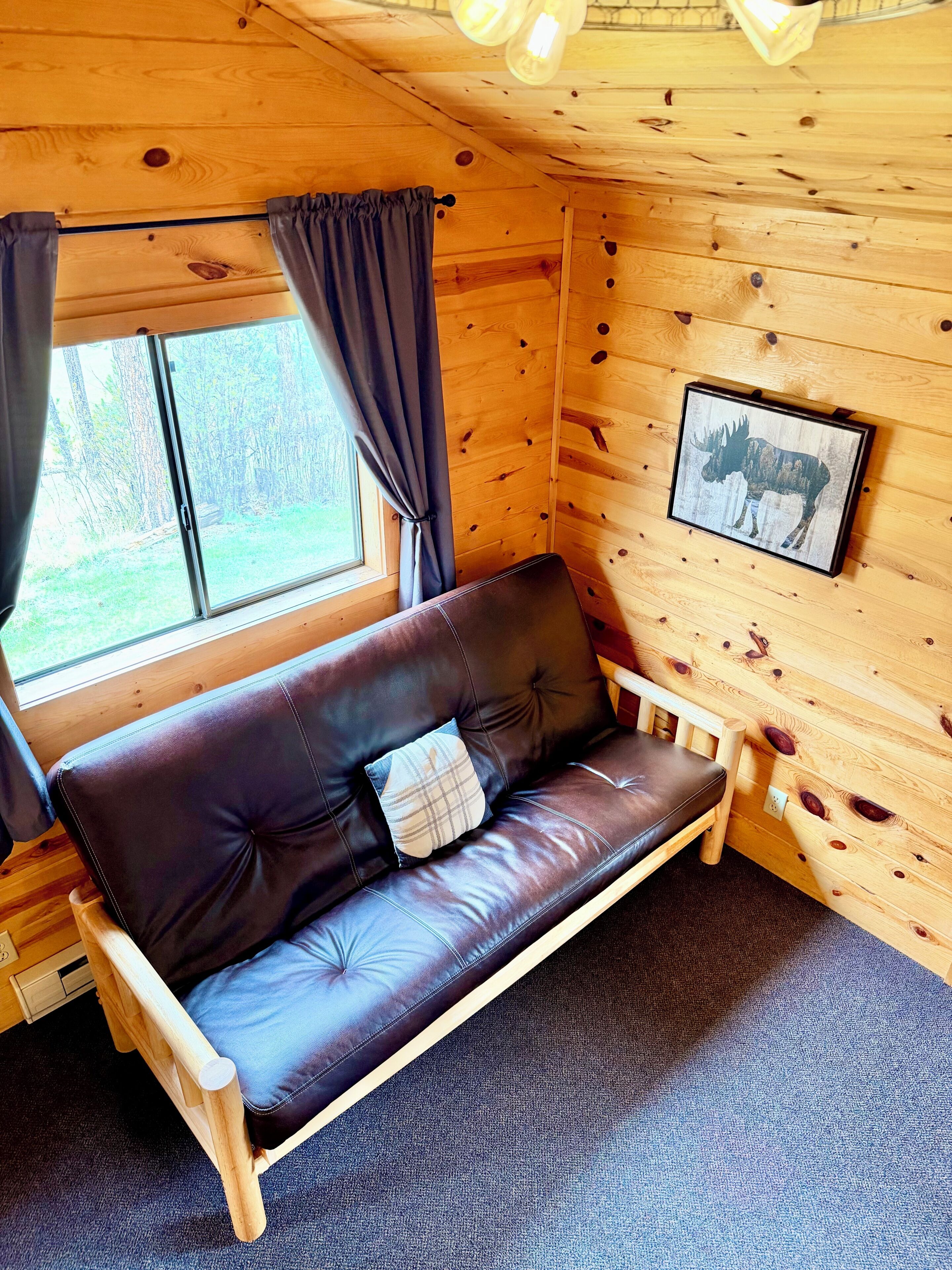 A wooden cabin interior with a leather sofa, a window with curtains, and a framed picture on the wall.