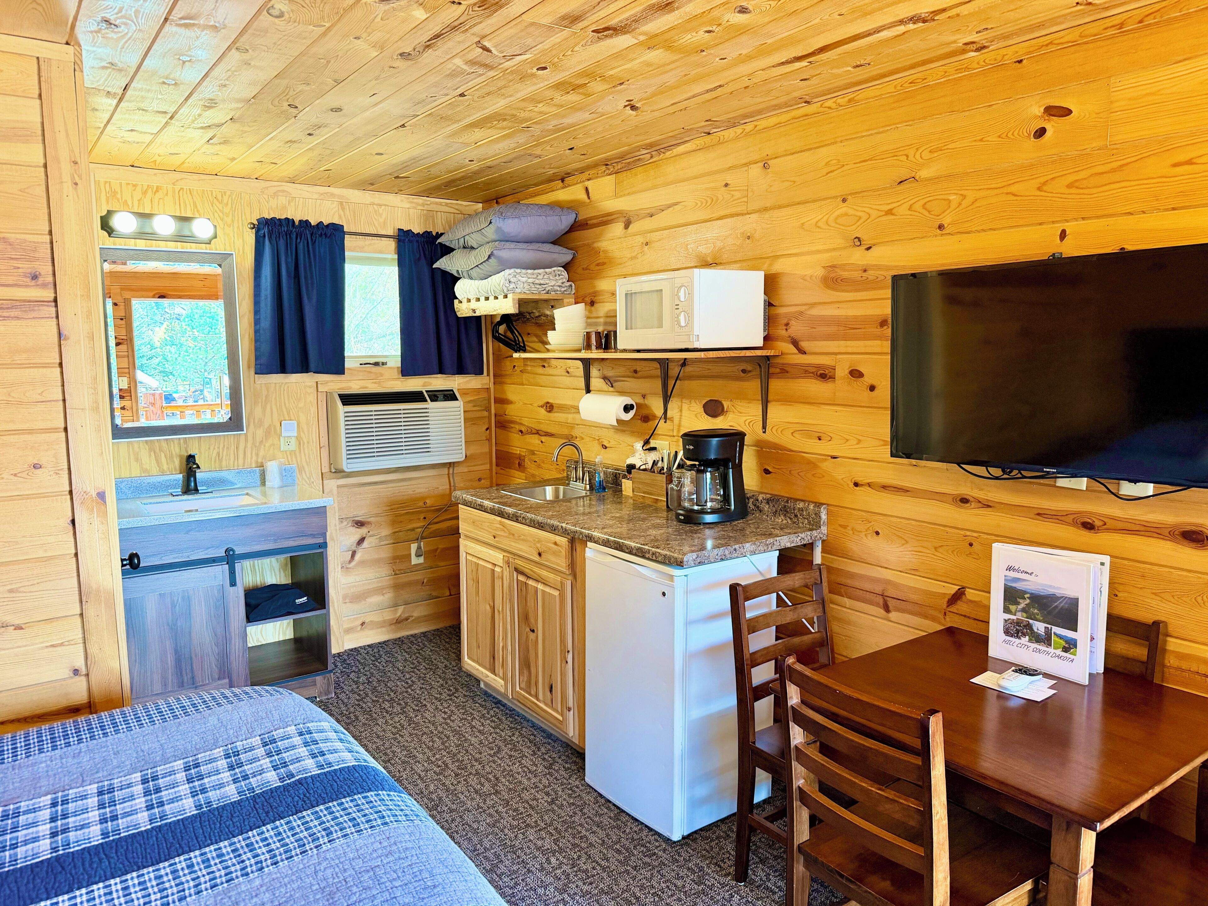 A compact cabin-style room with a kitchenette, a bed, a dining table, and a television.