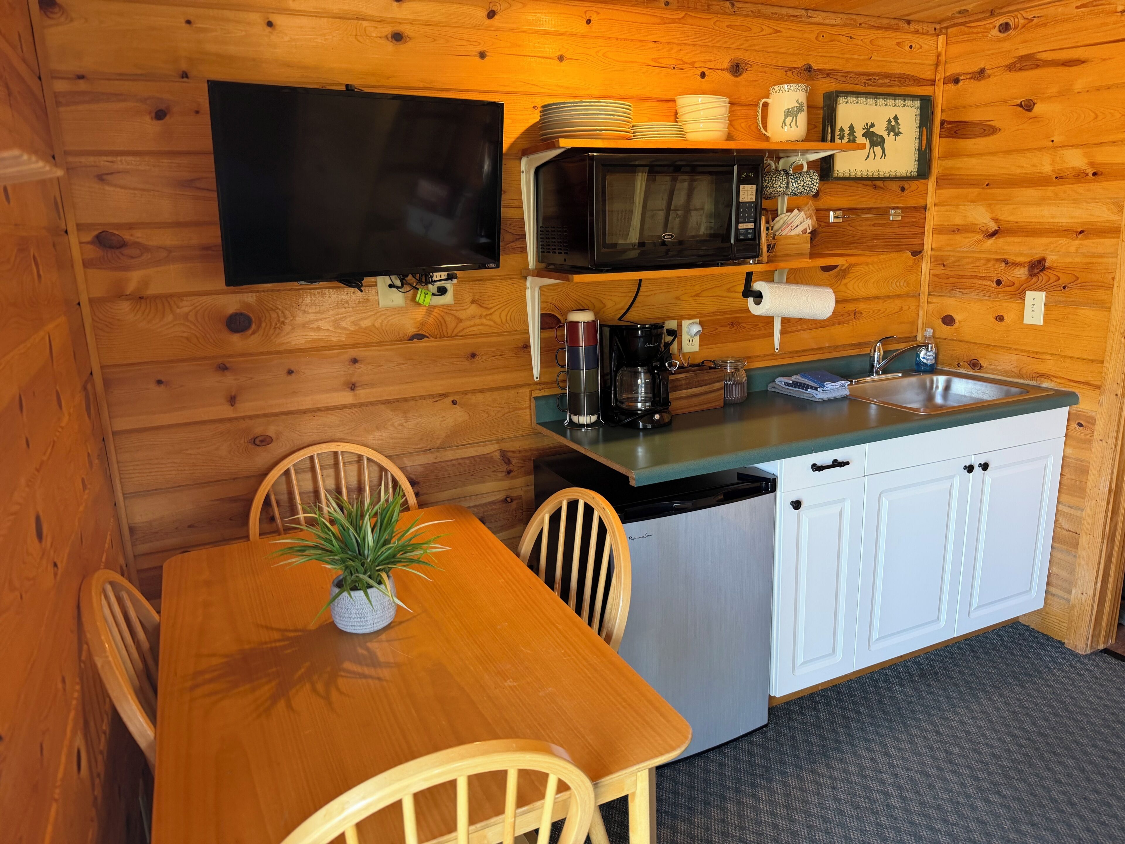 Una cocina compacta con paredes de madera, un televisor de pantalla plana, un microondas, un lavamanos y un comedor con sillas y una mesa.