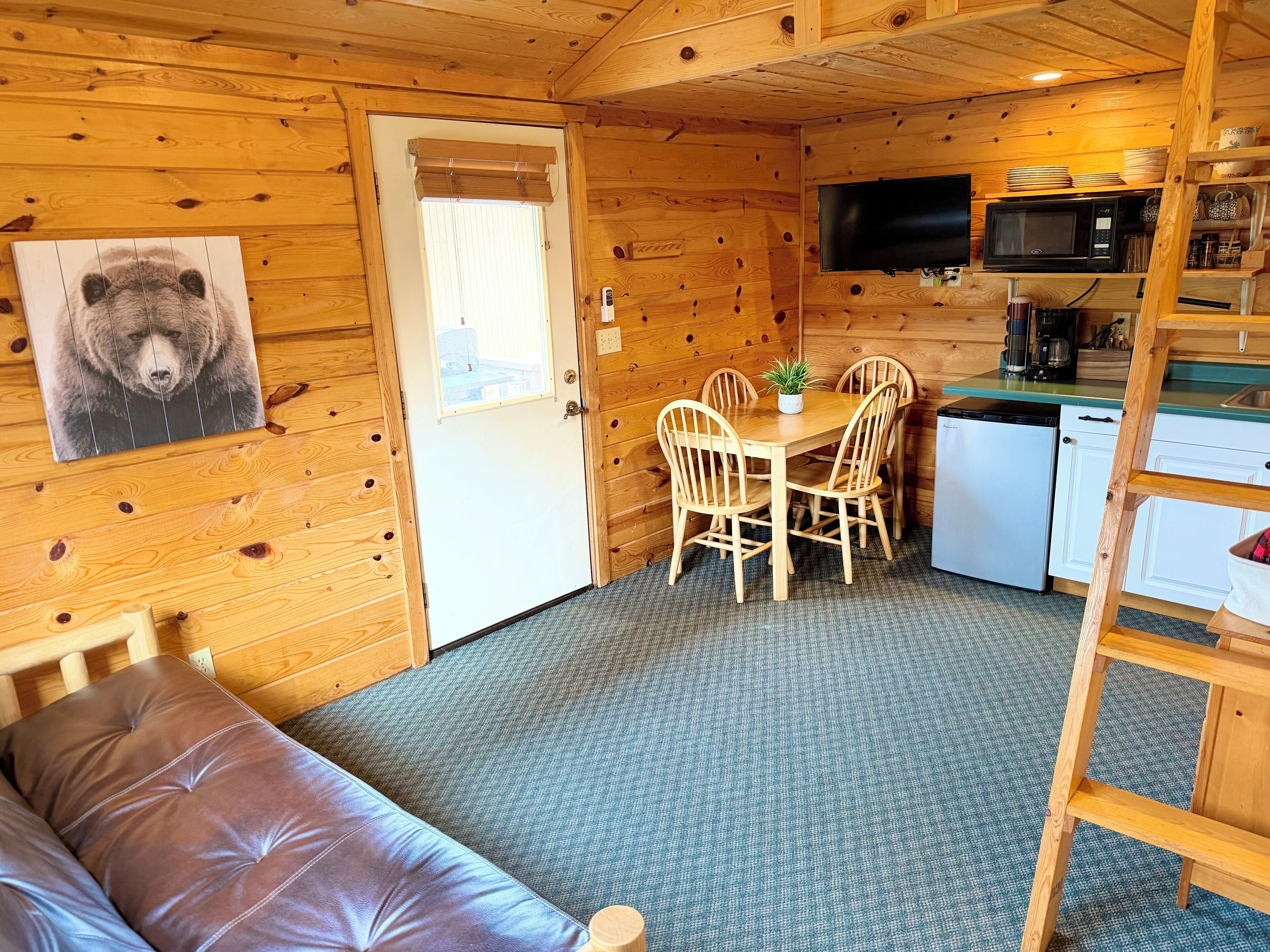 Interior de uma cabana de madeira com uma mesa de jantar, cadeiras, um sofá, cozinha com micro-ondas e televisão.