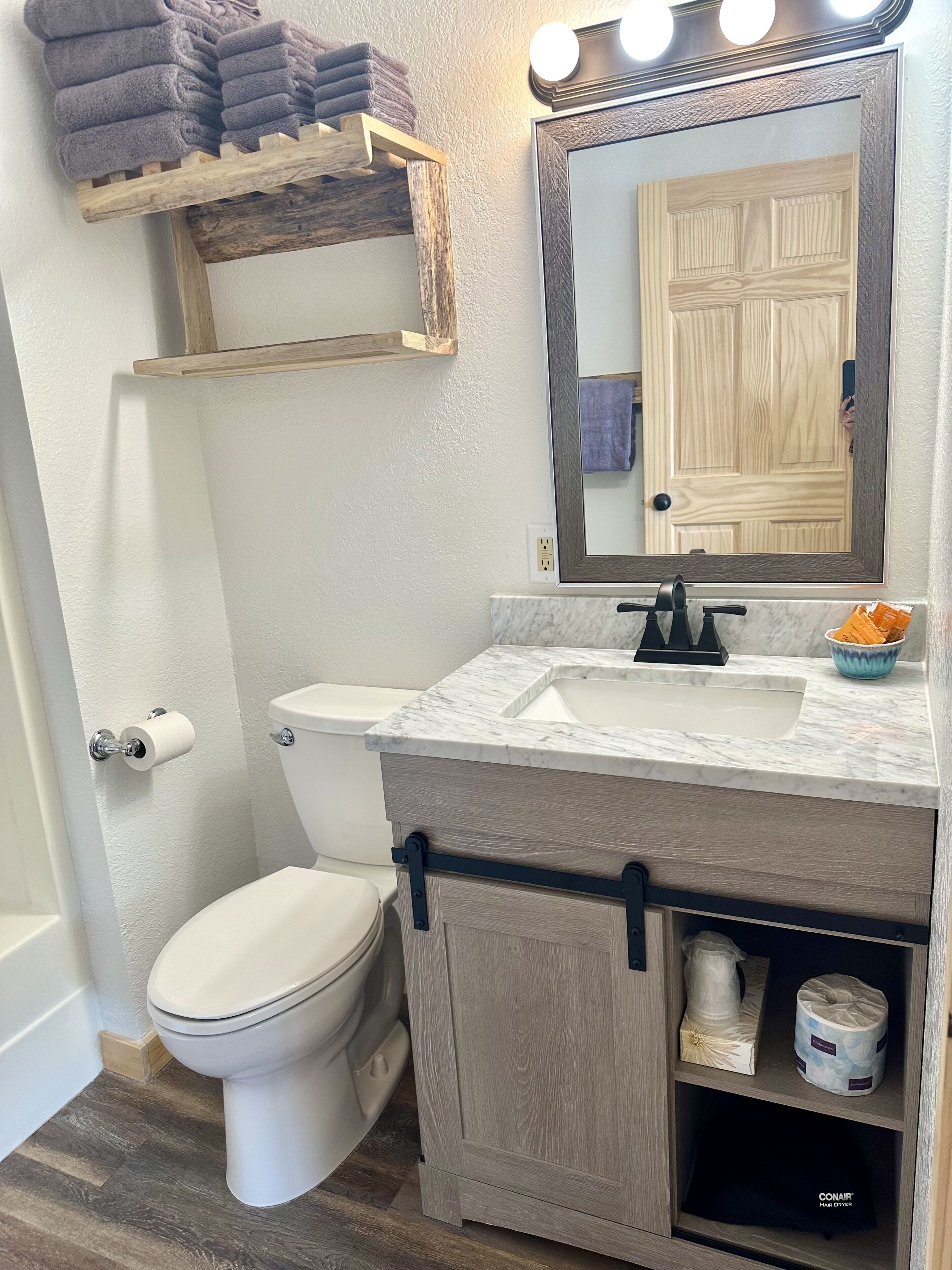 A bathroom with a toilet, sink, and mirror. There is a shelf with towels above the toilet and a hair dryer in the cabinet below the sink.