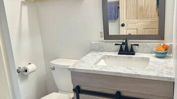 A bathroom with a toilet, sink, and mirror. There is a shelf with towels above the toilet and a hair dryer in the cabinet below the sink.