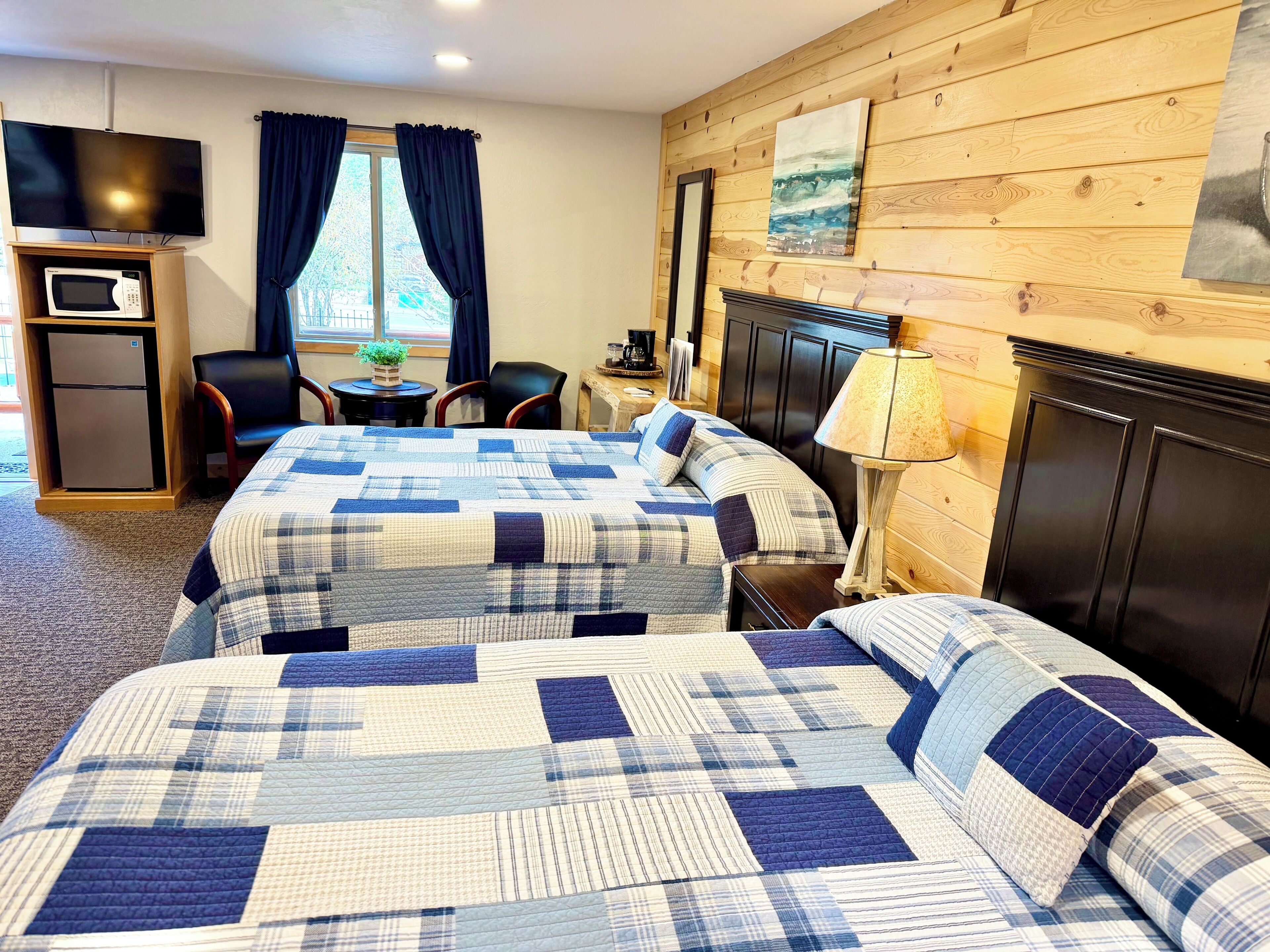 A hotel room with two beds, a TV, a small fridge, a desk with a lamp, and a window with curtains.
