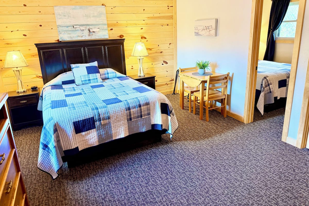 Une chambre à coucher avec un lit, des lampes de chevet, une tête de lit en bois, une petite table avec une plante et une porte donnant sur une autre pièce.