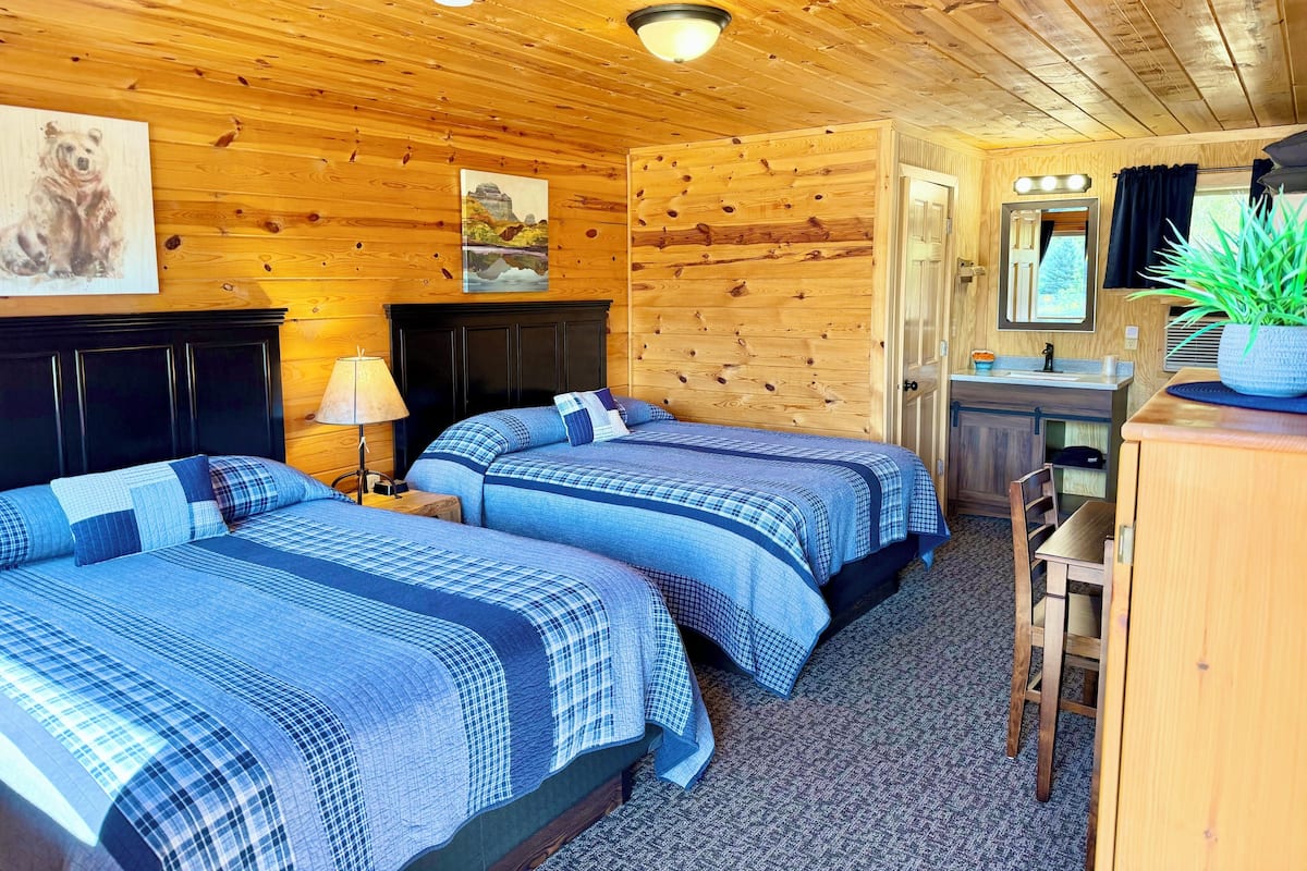 Une chambre de style cabane comprenant deux lits, un bureau, une chaise, une lampe, un miroir et une plante.