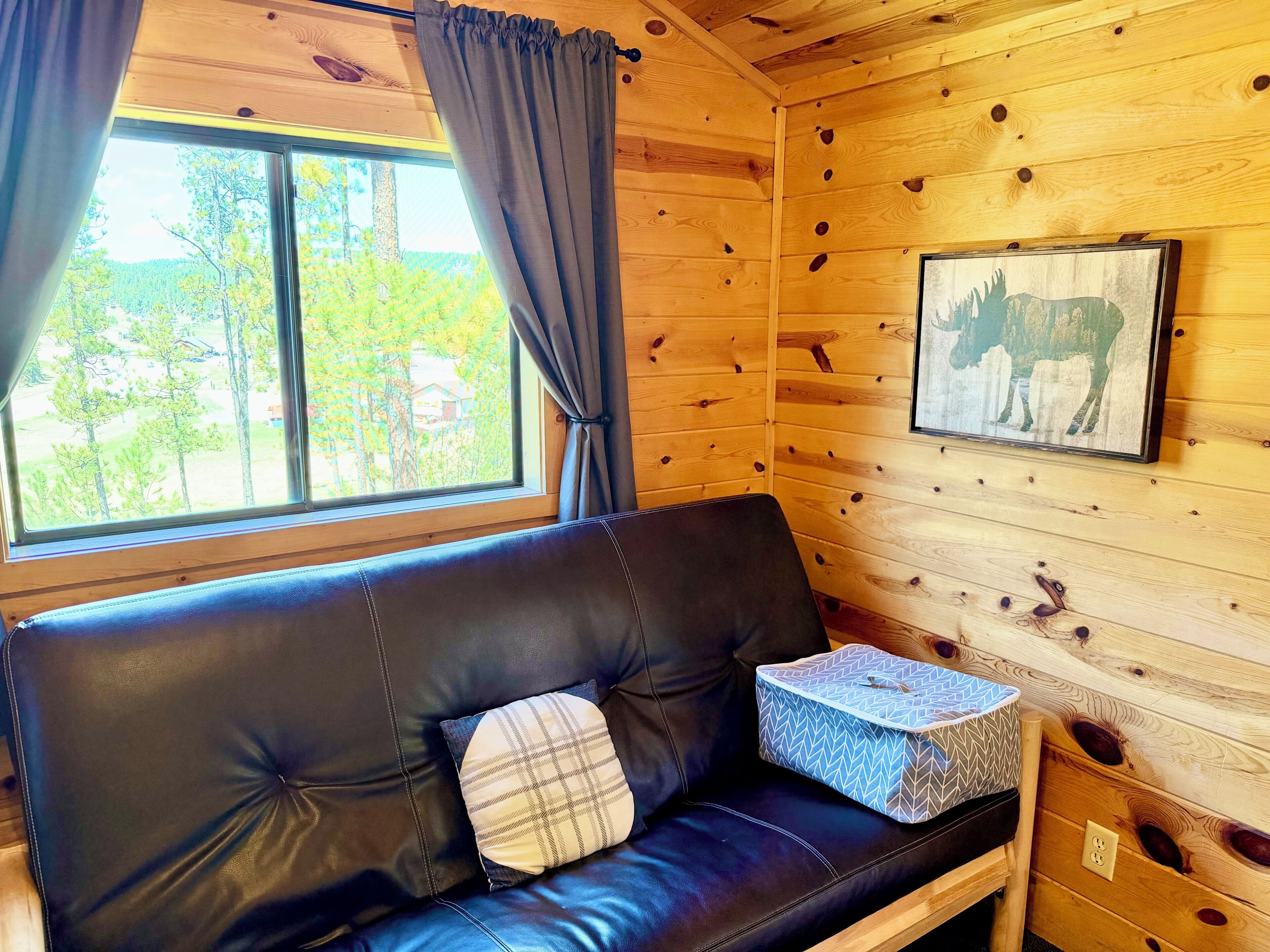 A wooden cabin interior with a black leather sofa, a plaid pillow, a framed picture of a moose, and a window with curtains.
