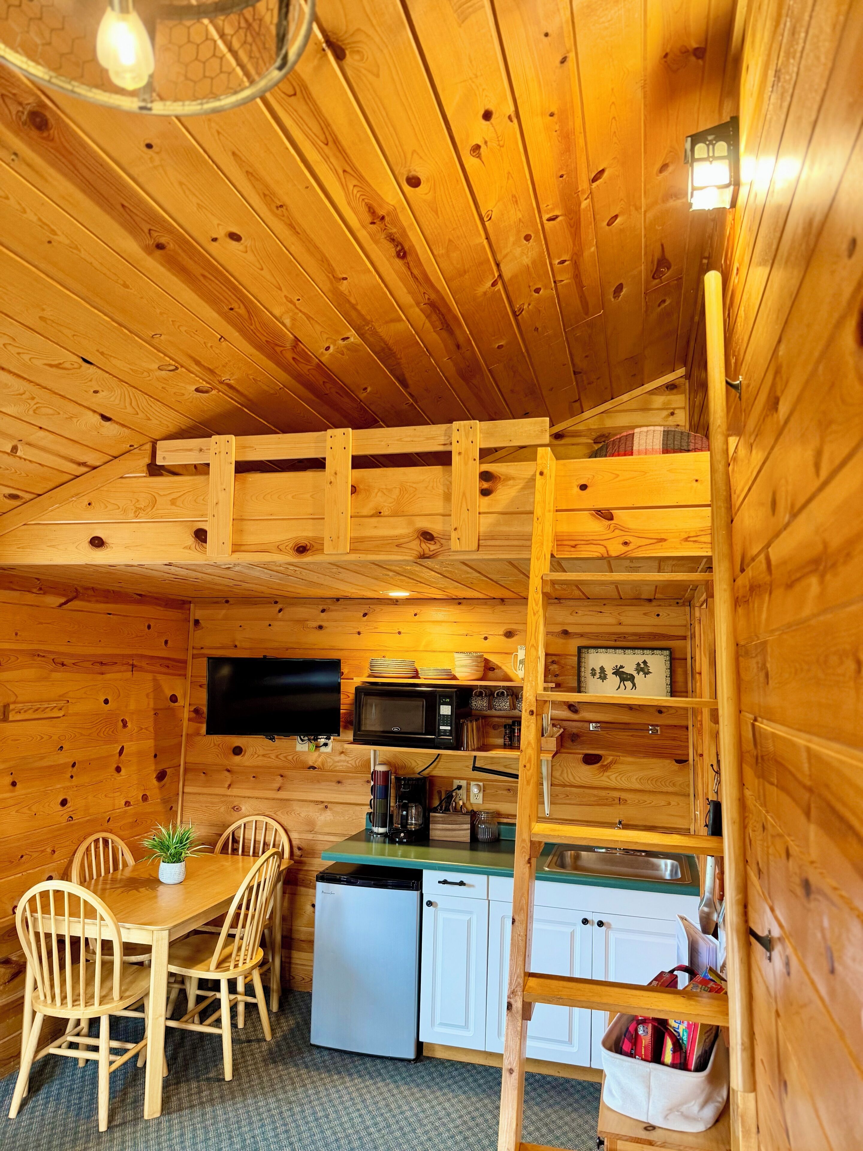 A compact kitchen with wooden walls, a dining table with chairs, a refrigerator, a microwave, and a small sink.