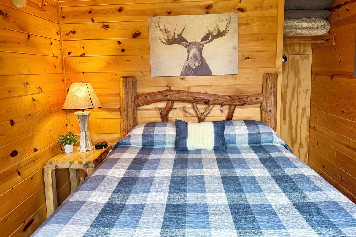 Une pièce de style cabane en bois, avec un lit, une table de chevet dotée d’une lampe, un tableau représentant une tête de cerf accroché au mur, et une petite plante.