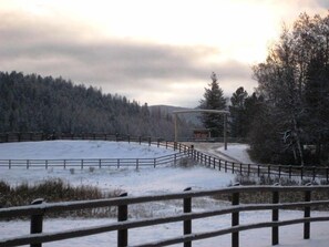 A snow-covered landscape with a fenced area and a wooden structure, likely a viewing platform or information booth, surrounded by trees and a distant mountain range. - Bar W Guest Ranch (Whitefish)