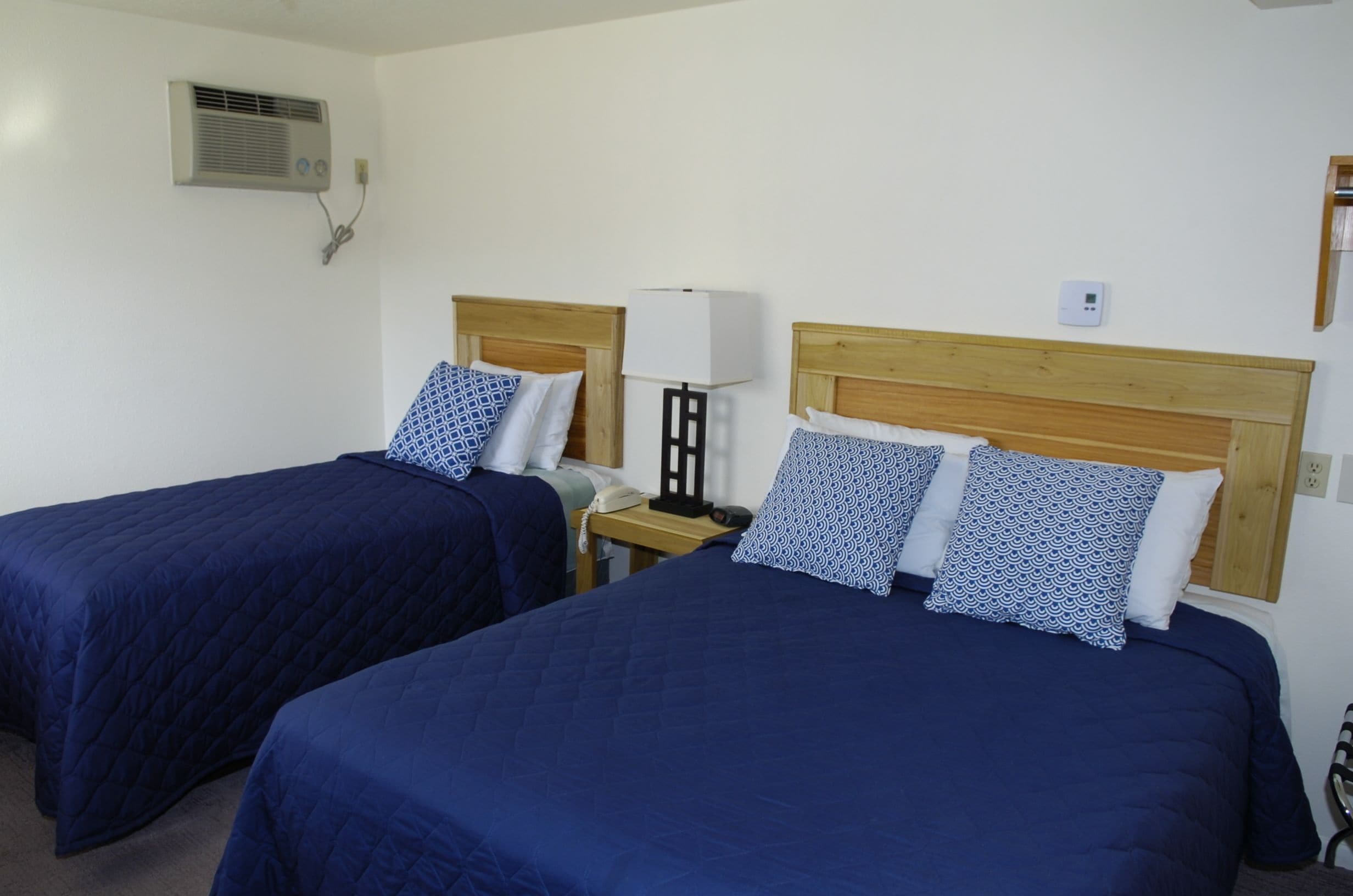Two single beds with matching headboards and blue bedding, a bedside table with a lamp, and a wall-mounted air conditioner.
