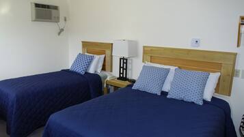 Two single beds with matching headboards and blue bedding, a bedside table with a lamp, and a wall-mounted air conditioner.