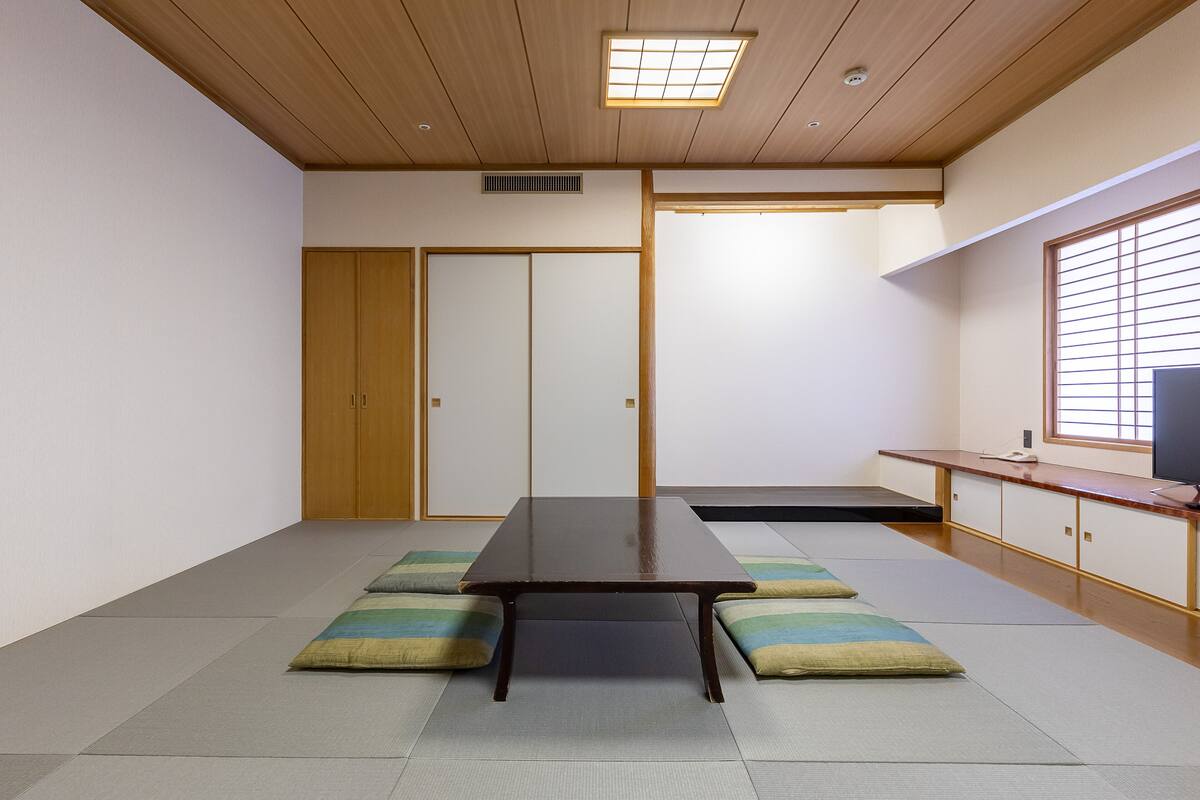 Une pièce de style japonais traditionnel, avec un sol recouvert de tatamis, une basse table en bois, des portes coulissantes et une vue sur l’extérieur à travers une fenêtre munie de stores.