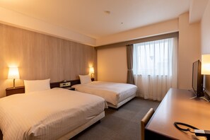 Ein Hotelzimmer mit zwei Betten, einem Schreibtisch und einem Stuhl. Nachttische mit Lampen befinden sich an beiden Seiten der Betten, und auf dem Schreibtisch steht ein Fernseher.