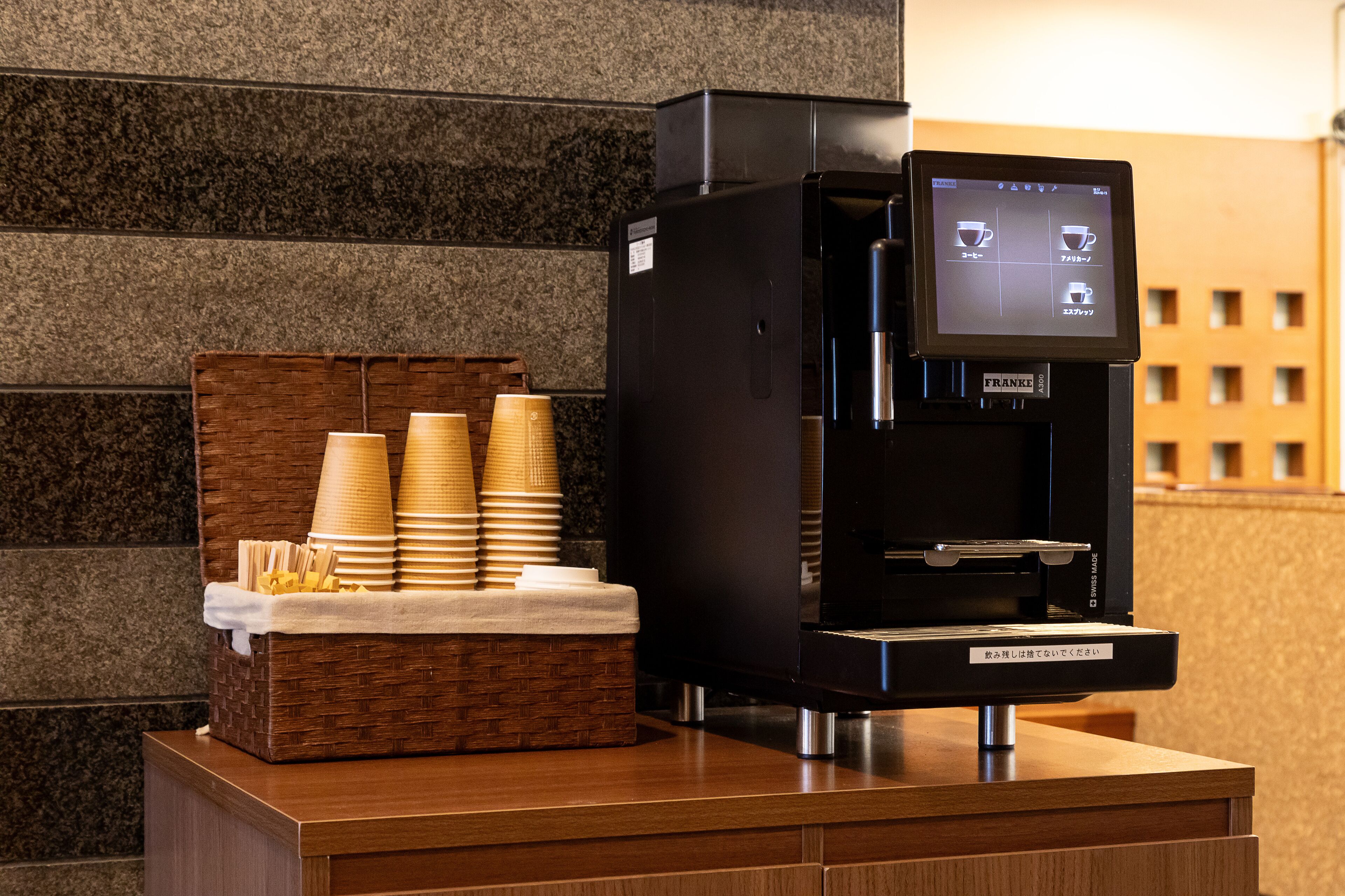 En kaffemaskine med digitalt display, stablede papirkrus og en kurv med servietter på en træbordplade.