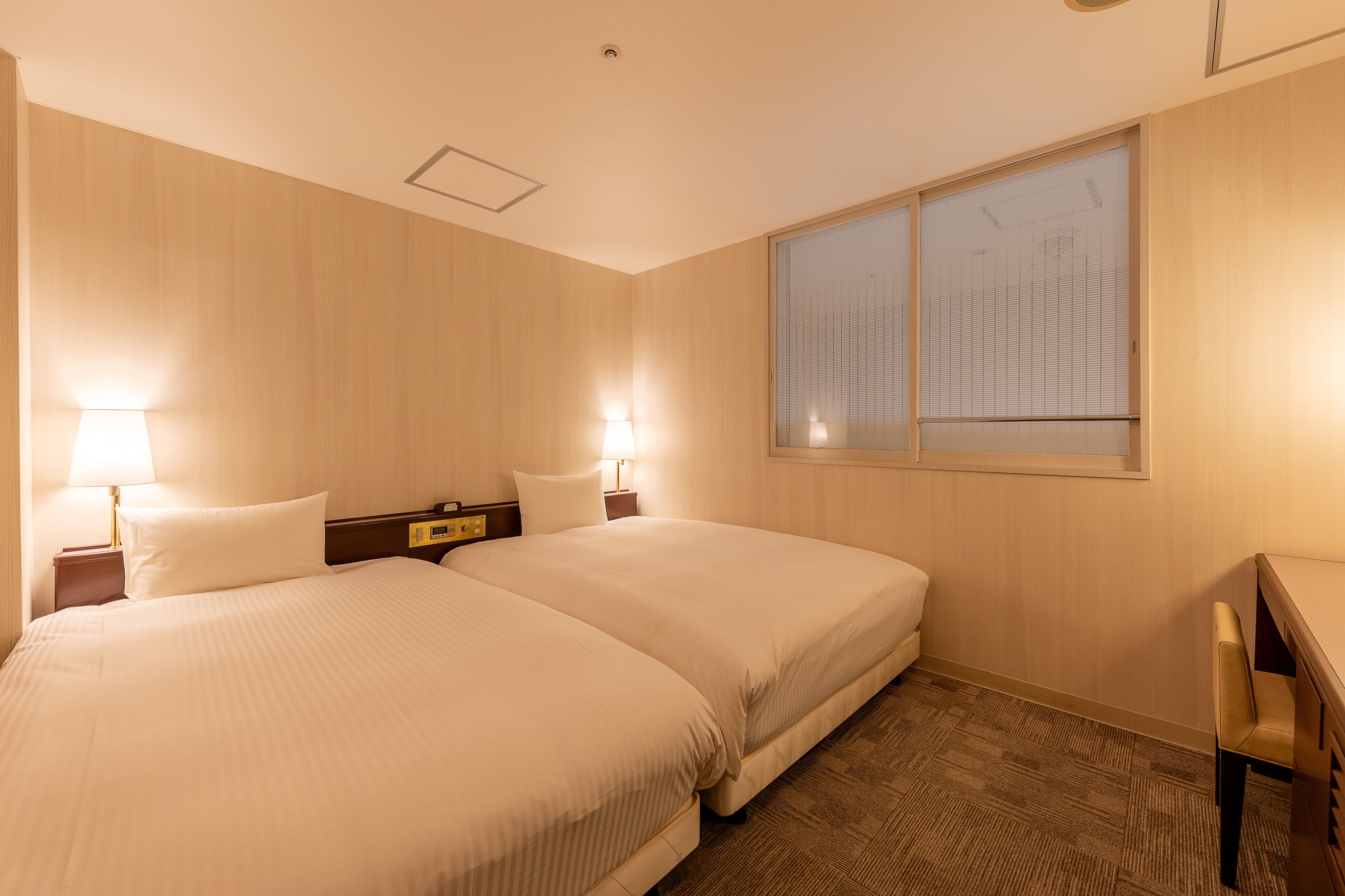 A hotel room with two beds, a nightstand with a lamp, a window with blinds, and a wooden desk with a chair.