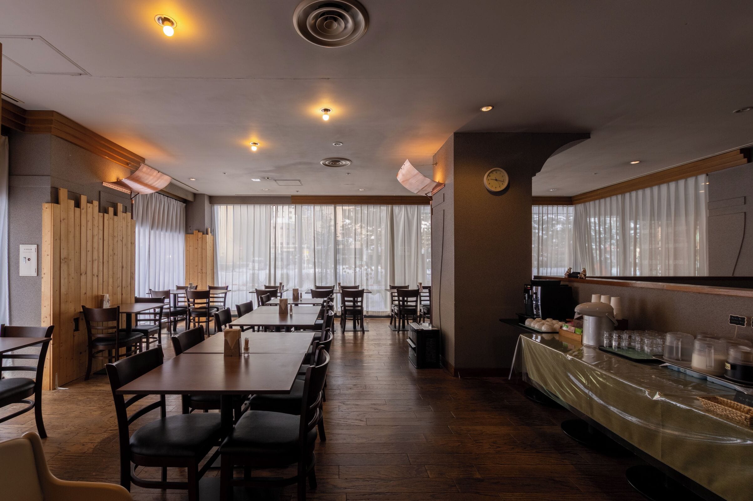 Un restaurant avec des tables et des chaises en bois, un comptoir où sont exposés des plats, et de grandes fenêtres avec des rideaux.