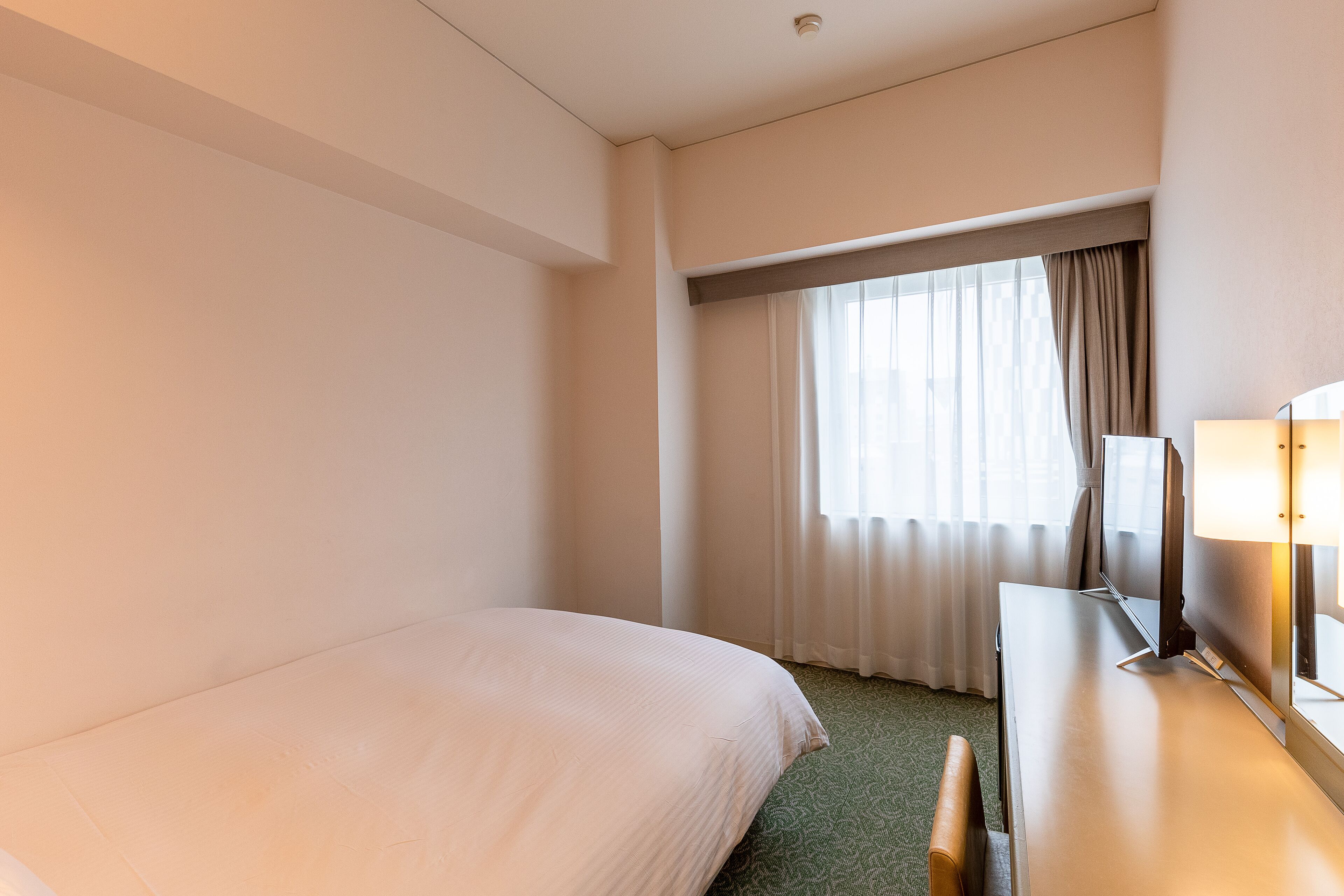 A hotel room with a bed, a desk with a lamp, and a window with curtains.