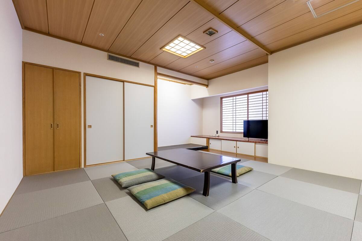 Une pièce de style japonais traditionnel, avec un sol recouvert de tatamis, une table basse, des coussins, des portes coulissantes et un téléviseur.