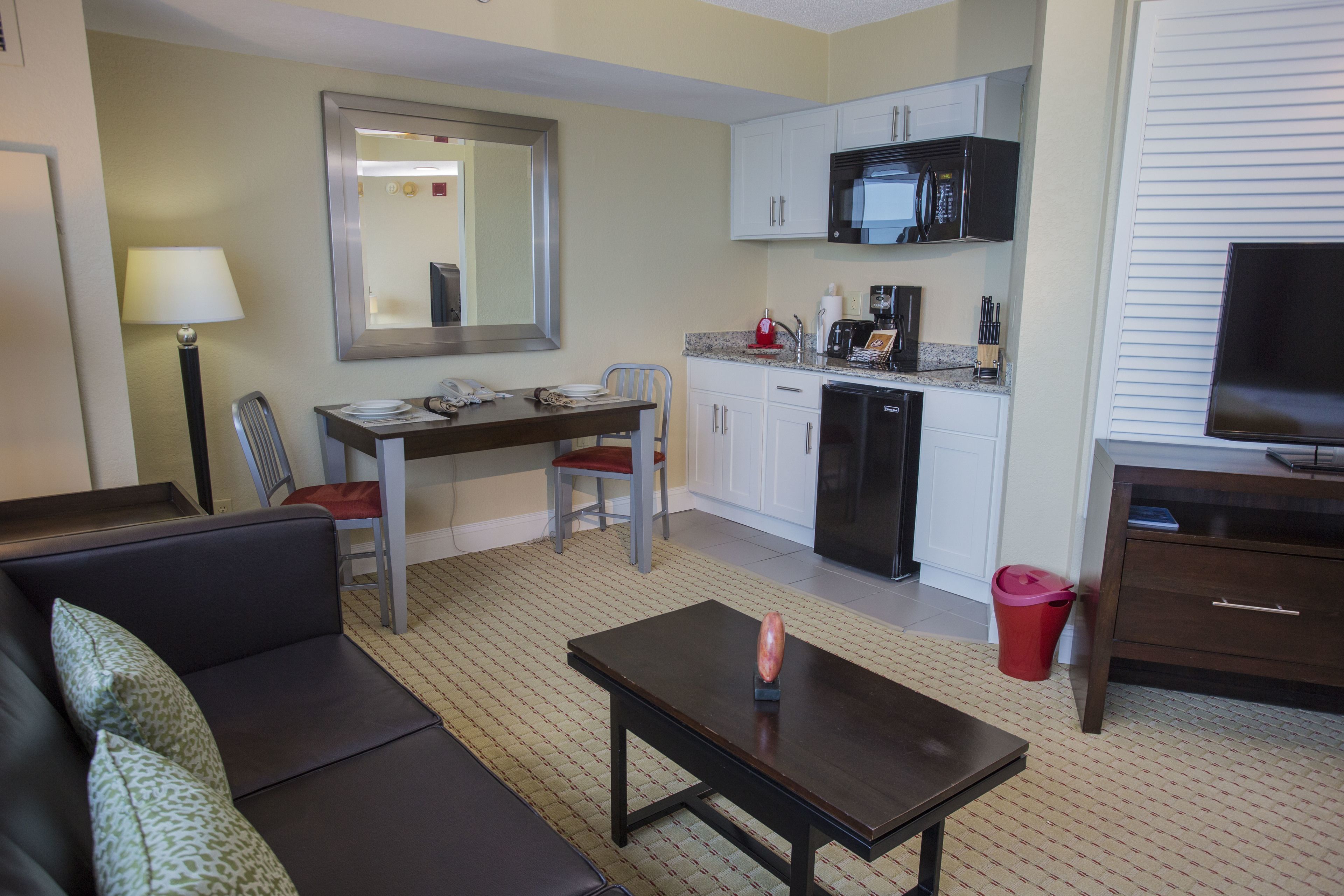 Habitación de hotel pequeña y funcional con sofá, una mesa de comedor pequeña con sillas, zona de cocina con microondas y televisor.