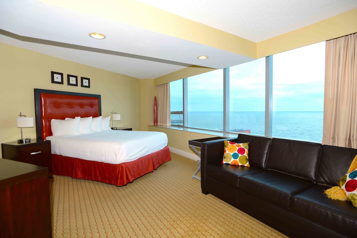 A hotel room with a large bed, a leather sofa, a desk, and a view of the ocean.