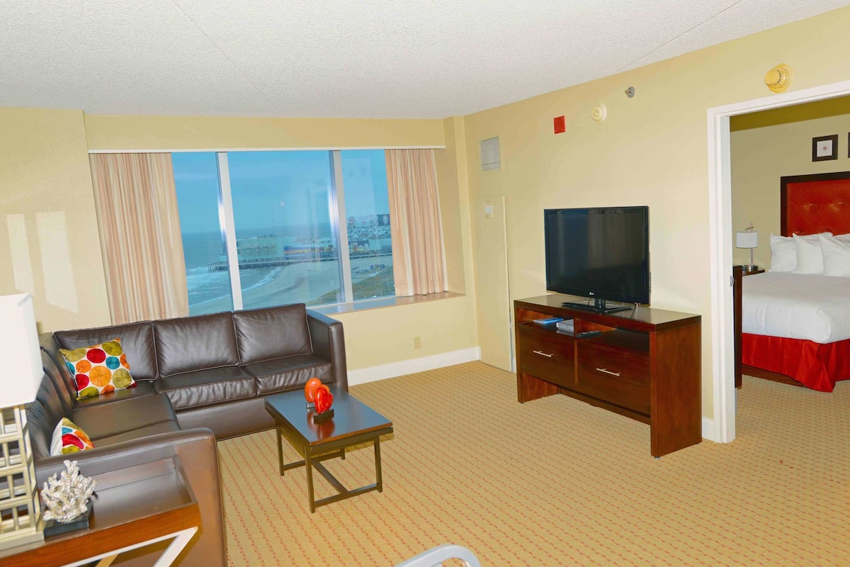 A hotel room with a large window, a television, a sofa, a desk, and a bed.
