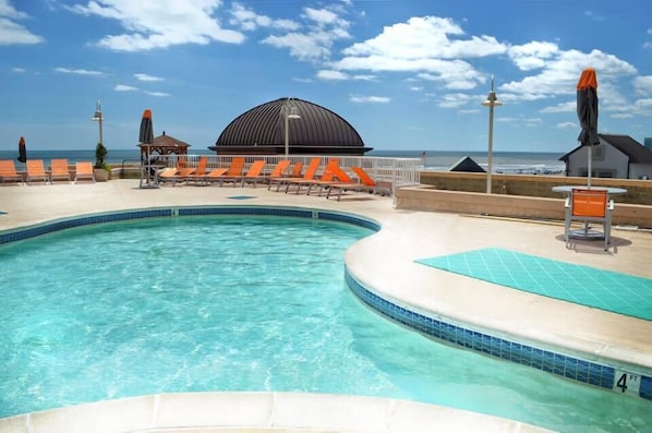 A pool area with lounge chairs and a covered seating area, a clear sky, and a view of the ocean. - Boardwalk Resorts Atlantic Palace (Atlantic City)