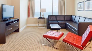 Une chambre d’hôtel moderne avec un grand canapé d’angle, un téléviseur à écran plat, une petite table et un pouf rouge.