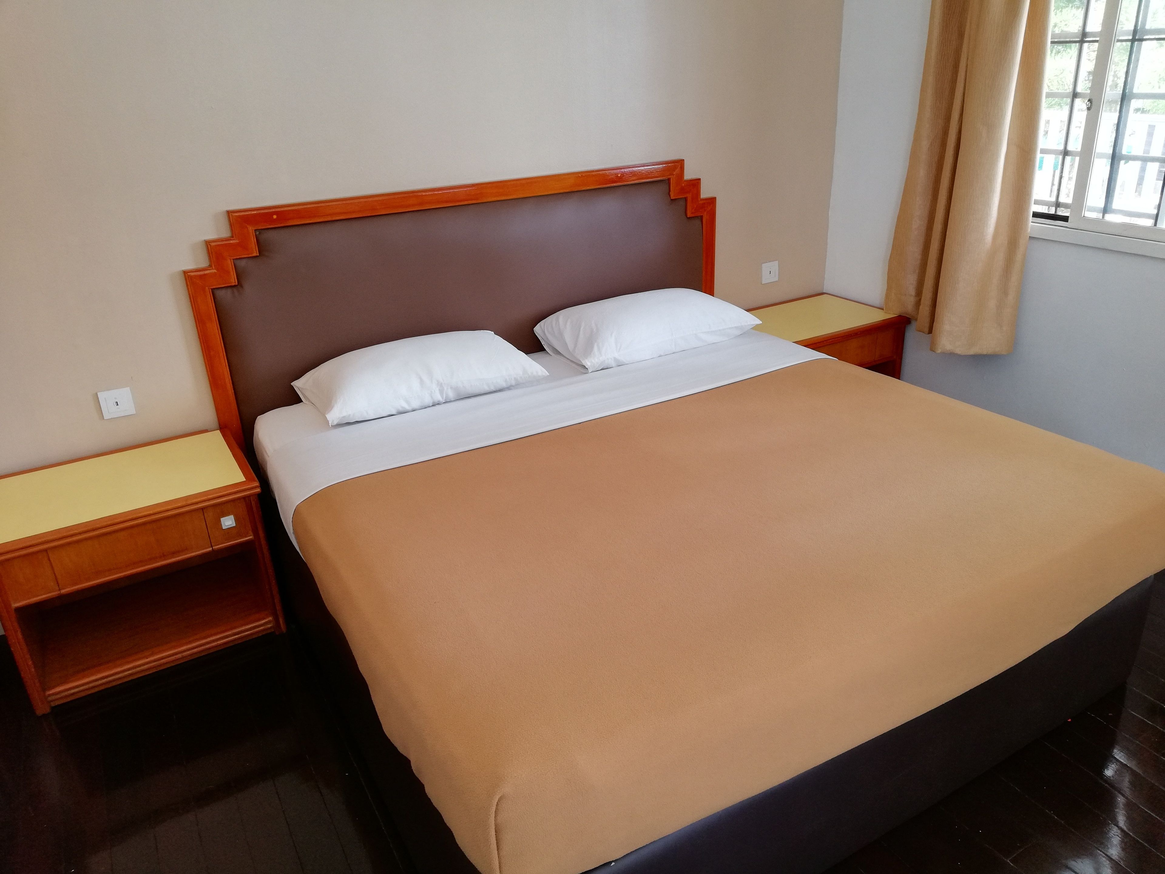 A neatly made bed with a brown bedspread and two white pillows, flanked by wooden nightstands with drawers, in a room with a window and a curtain.
