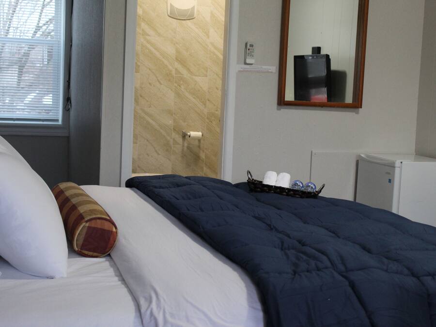 Una habitación de hotel con cama, espejo, refrigerador pequeño y ventana con persianas.