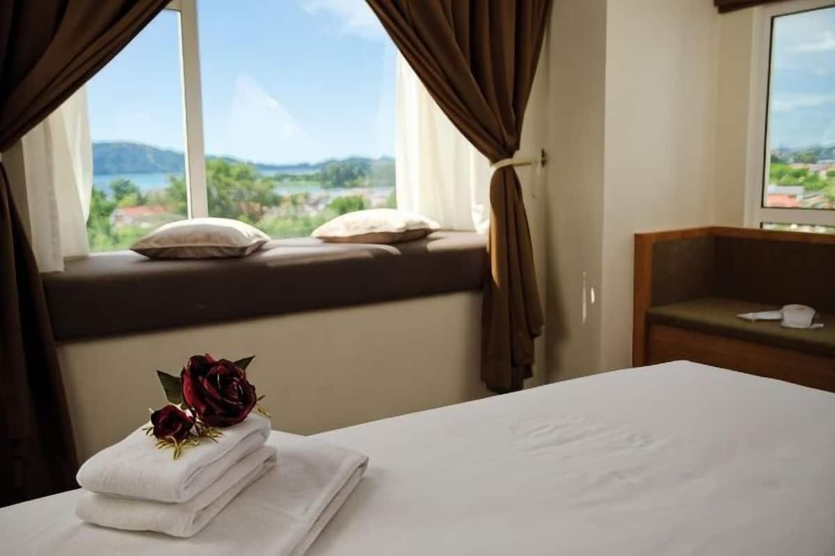 A hotel room with a bed, a window with curtains, a view of the outdoors, and a bedside table with a pillow.