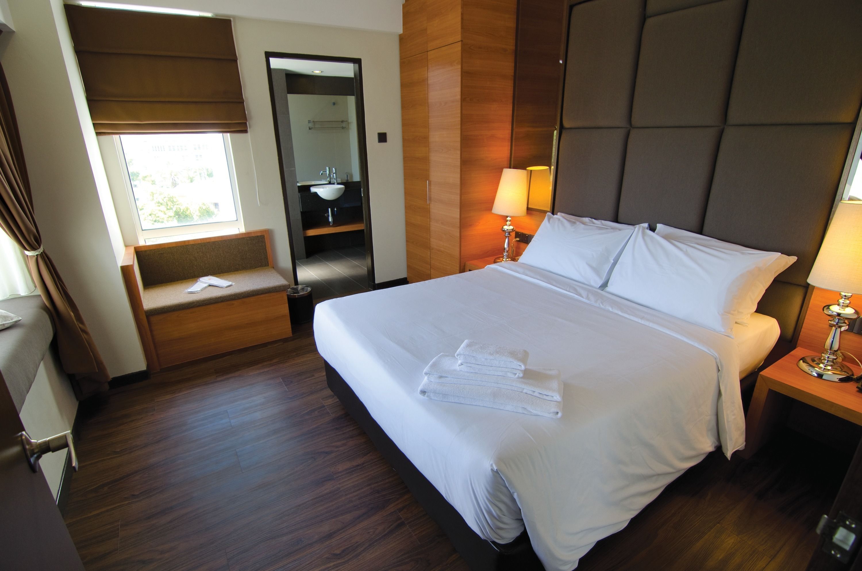 A hotel room with a large bed, a bedside table with a lamp, a window with a view, and a built-in bench with a mirror.