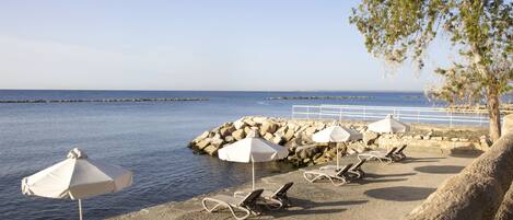 A beachside area with lounge chairs and umbrellas, a rocky shoreline, and a clear view of the sea.
