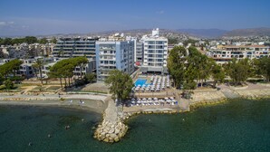 An aerial view of a coastal area with a swimming pool surrounded by beach chairs and umbrellas, a rocky shoreline, and residential buildings in the background. - Harmony Bay Hotel (Limassol)