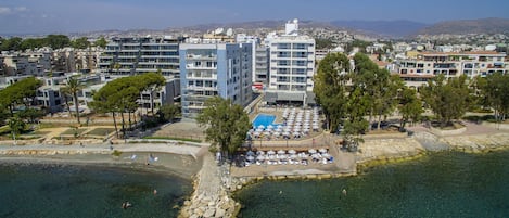 Vista aérea de una zona costera con piscina rodeada de sillas y sombrillas de playa, un litoral rocoso y edificios residenciales al fondo.