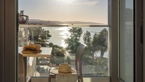 A balcony with a view of a body of water and distant hills, a chair, a small table with a hat, and a glass railing. - Harmony Bay Hotel (Limassol)
