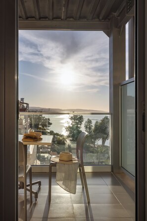 A balcony with a view of a body of water and distant hills, a chair, a small table with a hat, and a glass railing. - Harmony Bay Hotel (Limassol)
