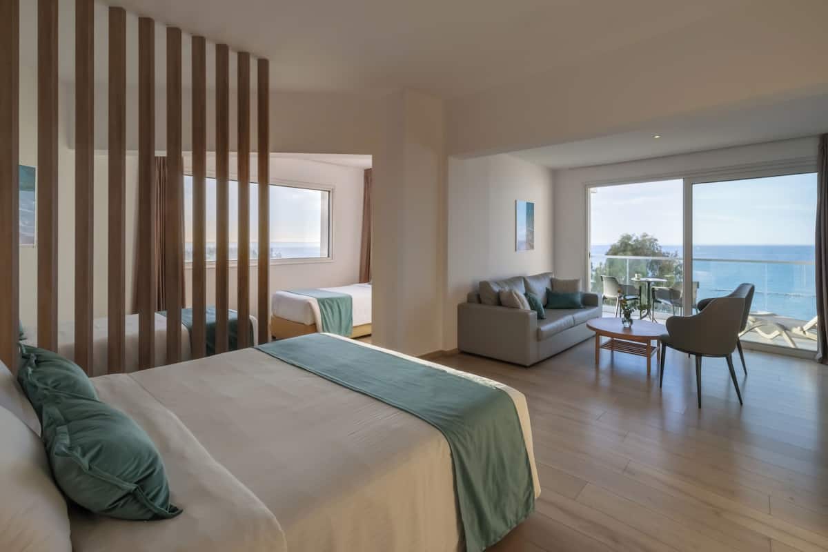 A hotel room with a large bed, a sofa, a dining table, and a balcony with a view of the sea.